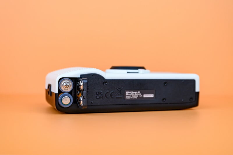 A close-up of the bottom of a white and black camera with a battery compartment open, exposing two AA batteries. The background is a solid orange color.