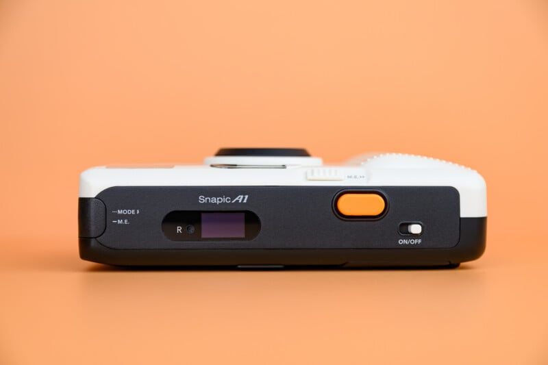 A compact point-and-shoot camera labeled "Snapic A1" displayed on its side against a solid peach background, showing its power button, mode switch, and control dial.