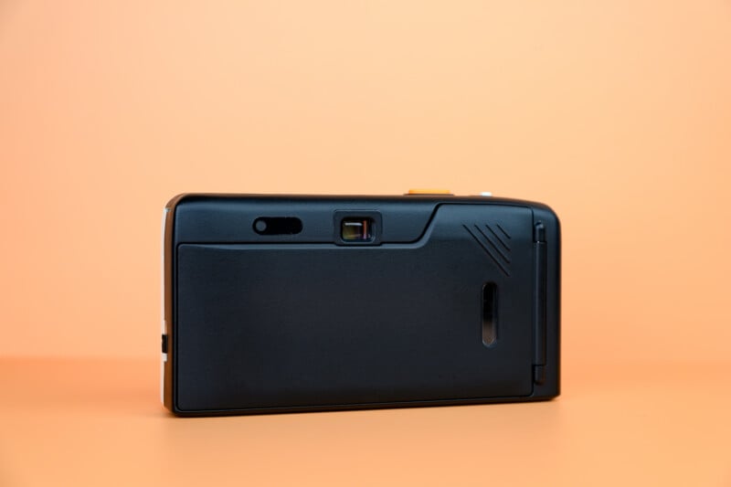 A black compact film camera is displayed against an orange background, shown from the back with its viewfinder, film door, and various camera details visible.