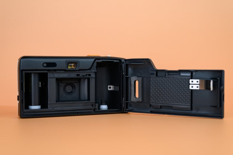 A black film camera with its back cover open, showing the film compartment and inner mechanisms, set against a plain peach-colored background.