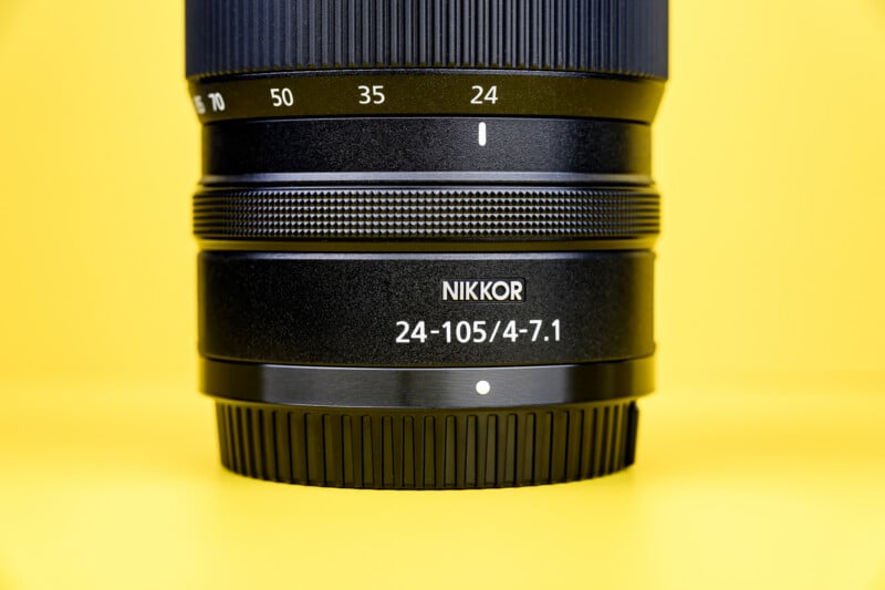Close-up of a NIKKOR camera lens with markings "24-105/4-7.1" against a bright yellow background. The lens details and focus ring are clearly visible.