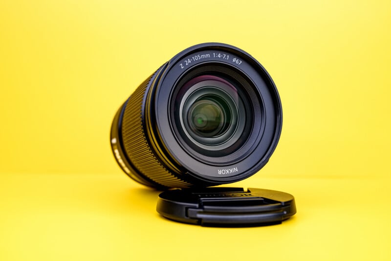 A Nikon camera lens with a 24-105mm focal length rests on its side with the lens cap placed underneath, set against a bright yellow background.