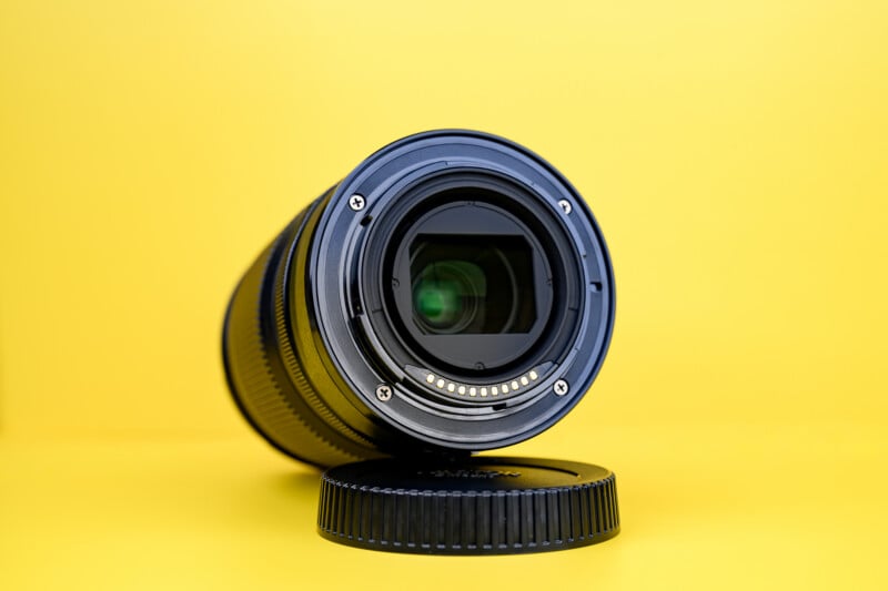 A close-up of a camera lens placed on its side with the lens mount facing forward, resting on a lens cap against a bright yellow background.