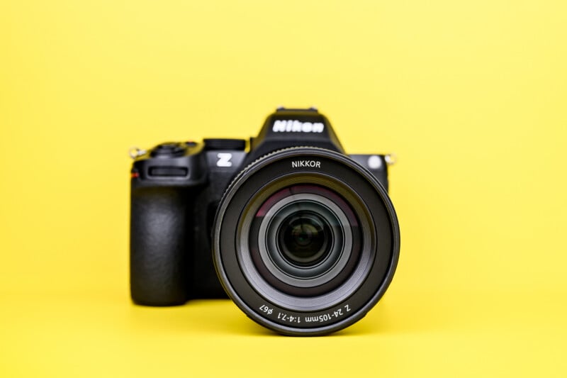 A Nikon Z series camera with a NIKKOR lens is positioned upright against a bright yellow background, facing directly forward.