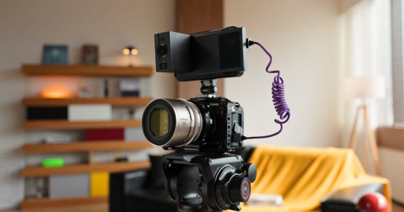 A professional camera with a large lens, external monitor, and coiled purple cable is mounted on a tripod indoors. A blurred background shows shelves, a yellow blanket, and a window with natural light.