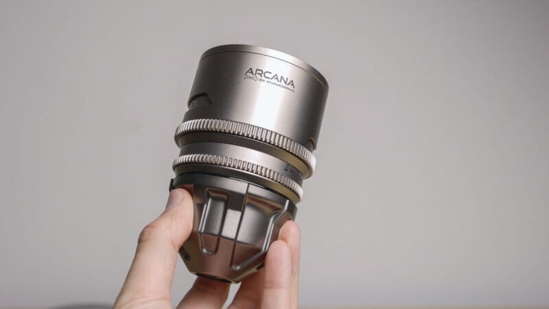 A hand holding a metallic ARCAN lens against a plain light gray background. The lens has a sleek, industrial design with gear-like rings and engraved branding near the top.
