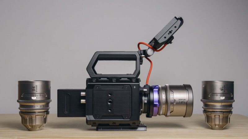 A professional cinema camera with a large lens is mounted on a wooden surface. Two additional camera lenses are positioned upright on either side of the camera. The background is plain and neutral.