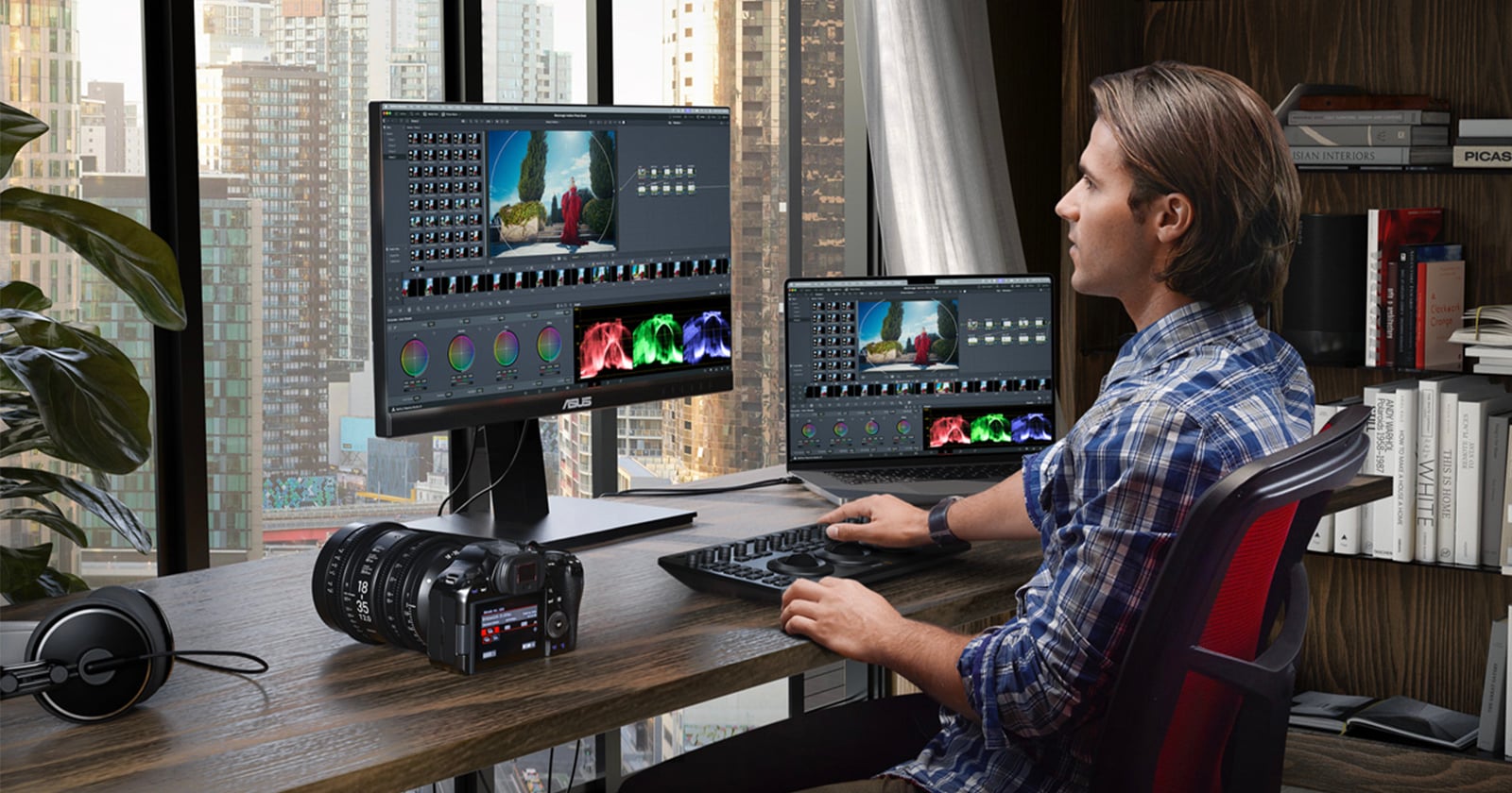 A man in a plaid shirt edits video footage on a desktop computer with two monitors in a modern office with large windows overlooking a cityscape. A camera, headphones, and books are on the desk.