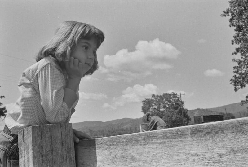 A young girl leans on a wooden fence, resting her chin on her hand and looking thoughtfully into the distance. Trees, clouds, and mountains are visible in the background under a clear sky.
