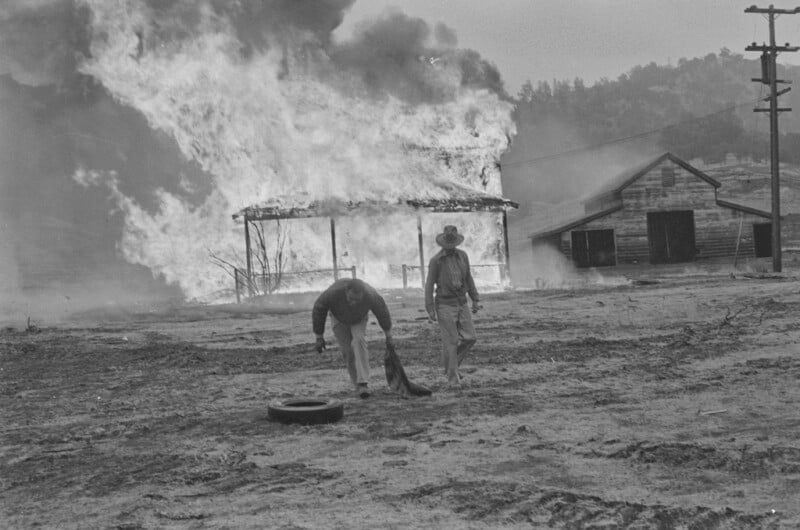 Two men walk away from a burning house engulfed in flames, with smoke rising into the sky. A wooden building stands nearby as the fire rages. The foreground is barren, and the mood is somber and tense.