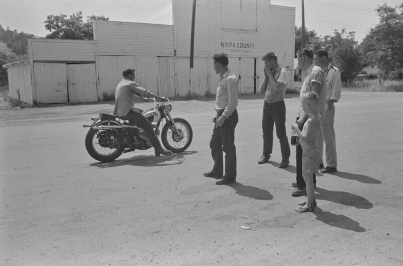 A man rides a motorcycle on a street while four men and a shirtless boy stand nearby watching. Behind them are white wooden buildings labeled "Napa County Districts." The scene appears to be from the mid-20th century.