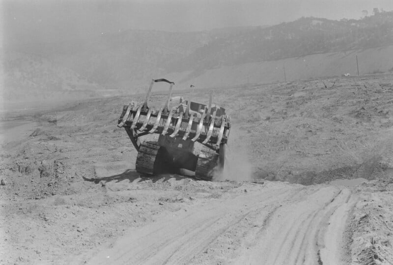 A bulldozer moves across a dusty, barren landscape, clearing earth. Tracks and ridges are visible in the foreground, with hills and a hazy sky in the background.