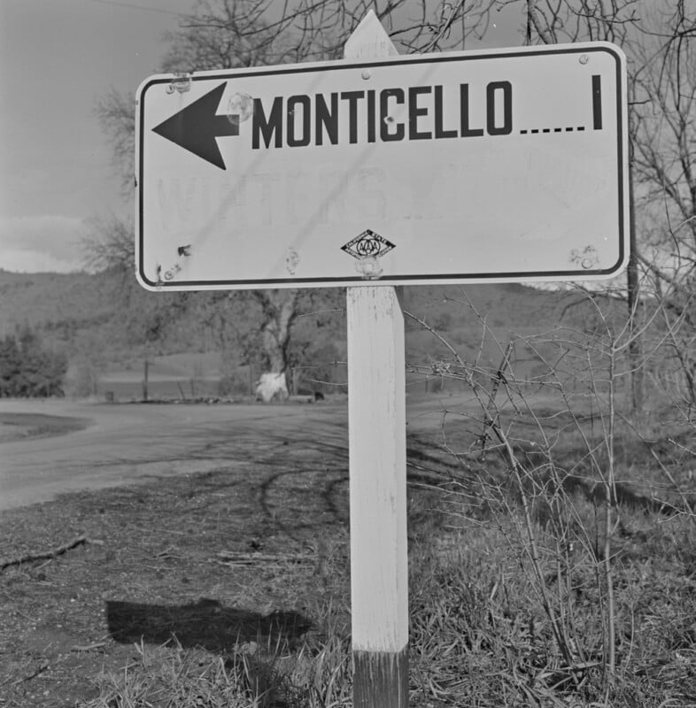 A road sign with an arrow pointing left reads "Monticello ..... 1" beside a rural road, with grass, bare trees, and hills in the background.