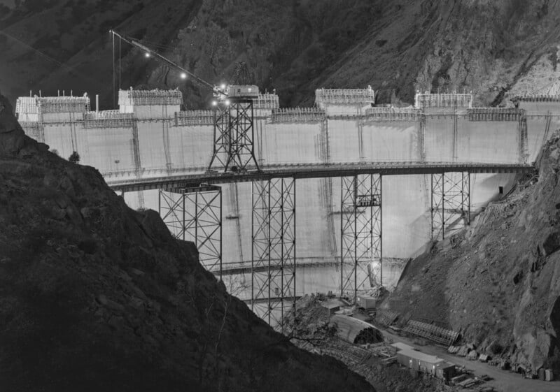 Black and white photo of the Hoover Dam under construction at night, illuminated by lights, with scaffolding and cranes in front of the partially finished concrete structure set among rocky hills.
