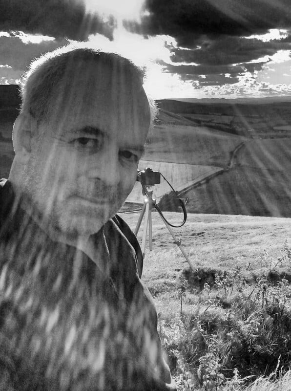 A man takes a selfie outdoors on a grassy hill under dramatic sunlight, with sunbeams streaming through clouds. A camera on a tripod is set up behind him, overlooking a rural landscape. The photo is in black and white.