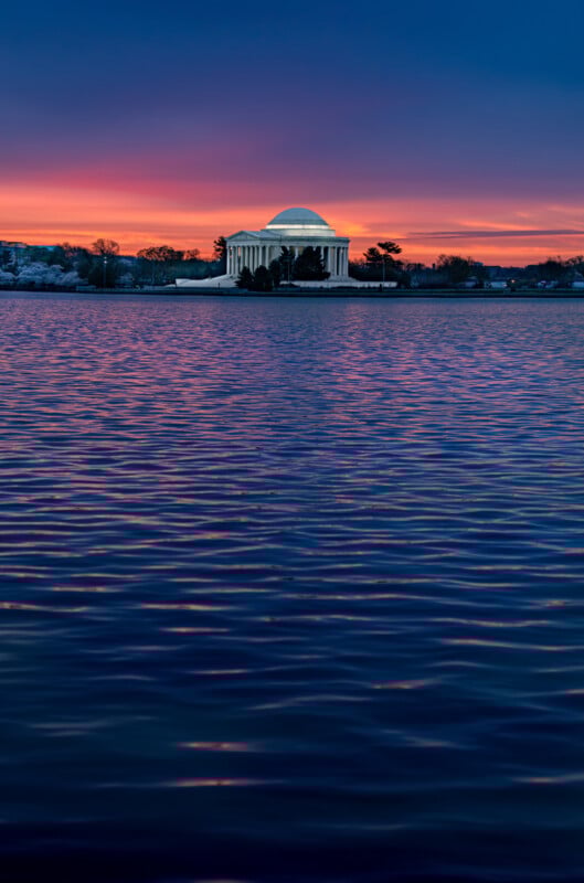 The Jefferson Memorial stands across rippling water at sunrise, with a vibrant sky blending deep blue, orange, and pink hues above the domed structure in Washington, D.C.