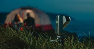 A camera on a tripod is set up in tall grass, focused on a blurred tent in the background with two people sitting inside, during dusk near a body of water.