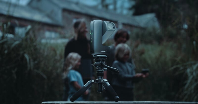 A compact camera on a tripod is in sharp focus outdoors, while four people, including two children, stand blurred in the background among tall grass.