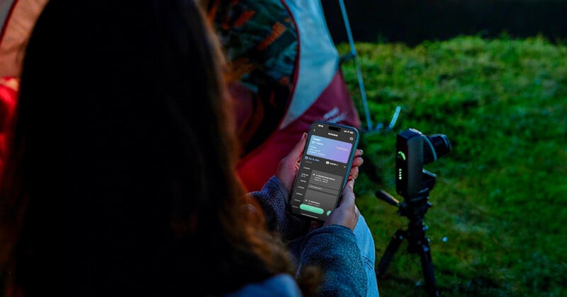 A person sitting outdoors near a tent holds a smartphone displaying an app, with a camera mounted on a tripod nearby and grass visible in the background.