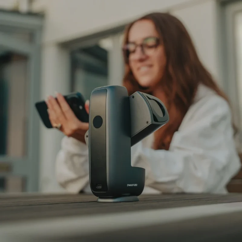 A woman with long hair and glasses holds a smartphone in the background, while a compact black device with the label "DWARF II" sits on a wooden table in the foreground.