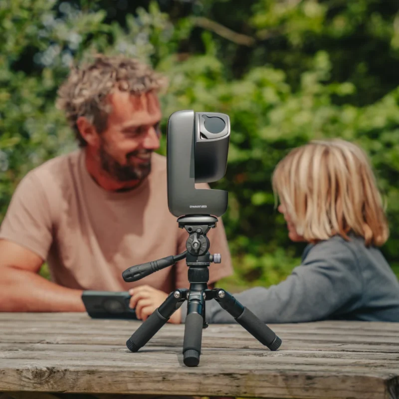 A man and a child sit at a wooden table outdoors, blurred in the background. In focus at the front is a camera mounted on a tripod, facing them, with green foliage visible behind.