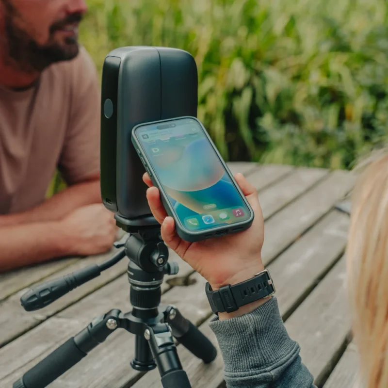A person holds a smartphone near a black electronic device mounted on a tripod, while another person sits nearby at a wooden outdoor table with greenery in the background.