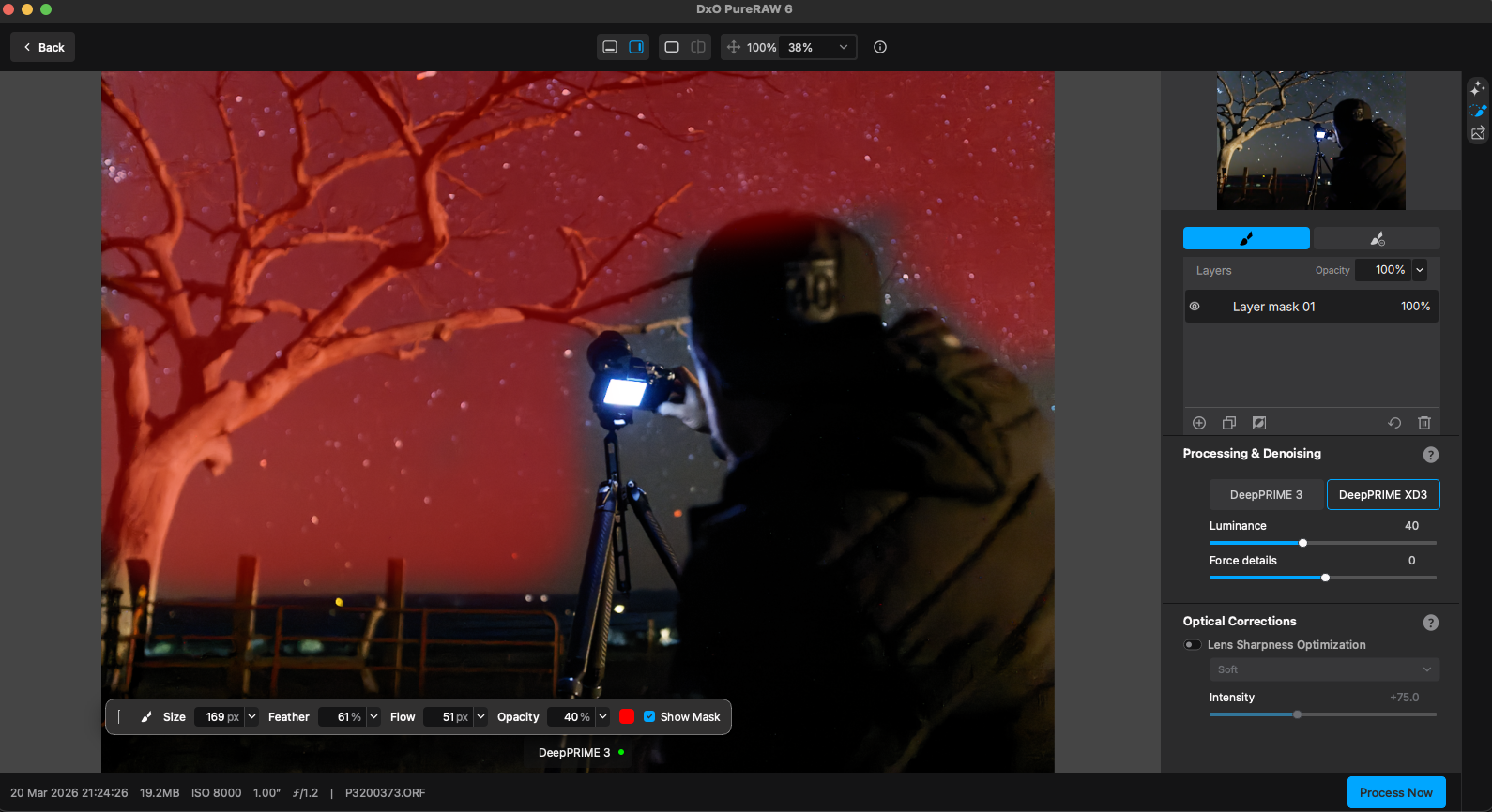 A person in dark clothing takes a photo of a vivid red night sky filled with stars, while editing the image on a computer screen using photo editing software. A leafless tree is visible on the left.