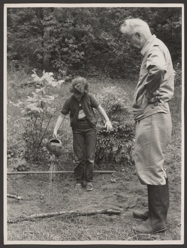 A young person waters a garden with a watering can while an older man with white hair and boots stands nearby, watching in an outdoor setting with trees and plants in the background.