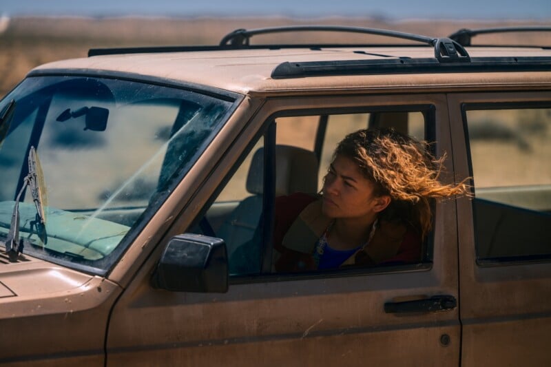A person with long hair blowing in the wind sits in the driver's seat of a dusty brown SUV, looking out the open window in a desert landscape.