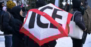 A group of people stand outside in winter clothing, with one person holding a red and white kite or sign that says "ICE" in large letters, crossed out with red tape. Snow is visible on the ground.