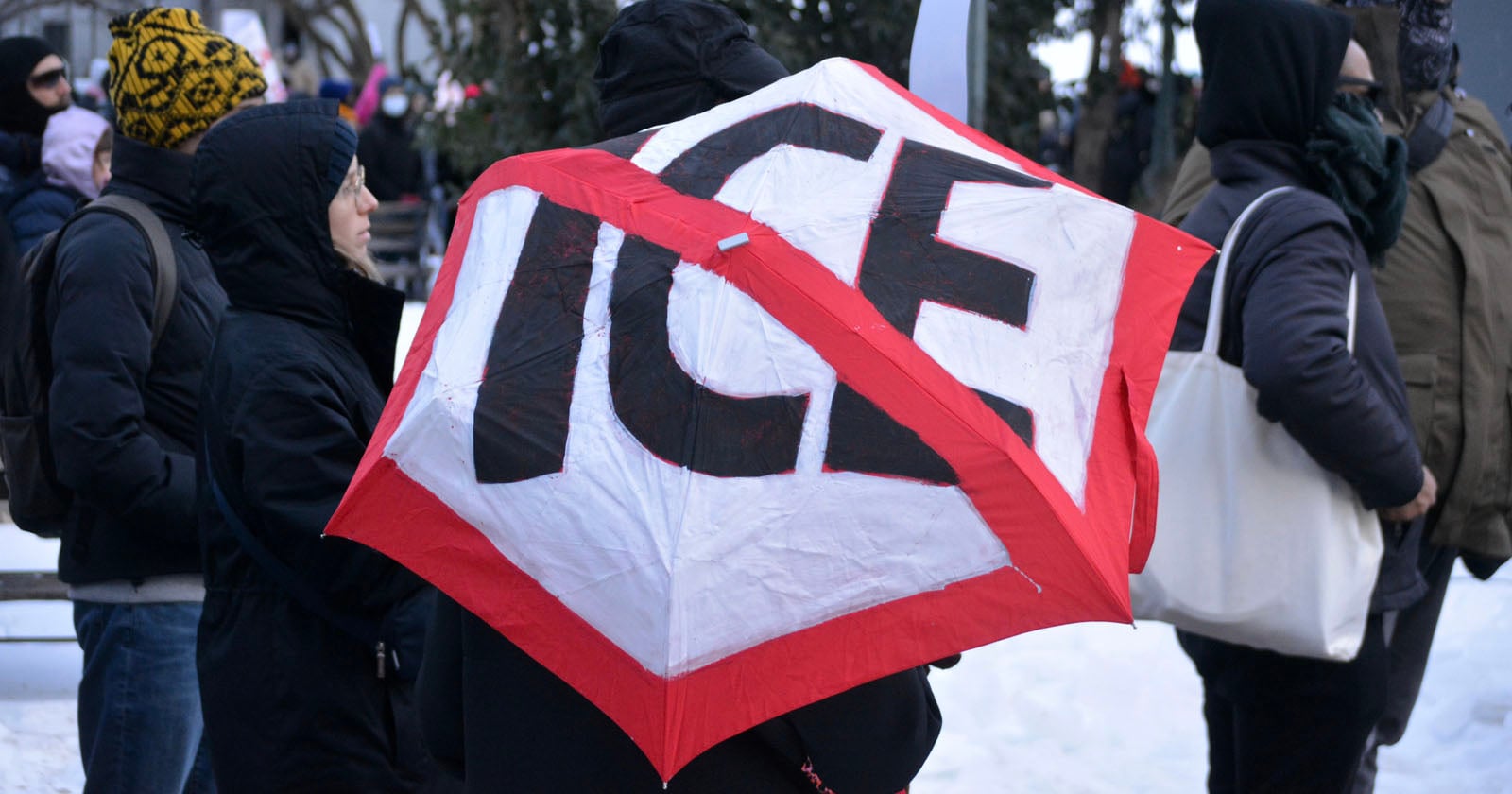 A group of people stand outside in winter clothing, with one person holding a red and white kite or sign that says "ICE" in large letters, crossed out with red tape. Snow is visible on the ground.