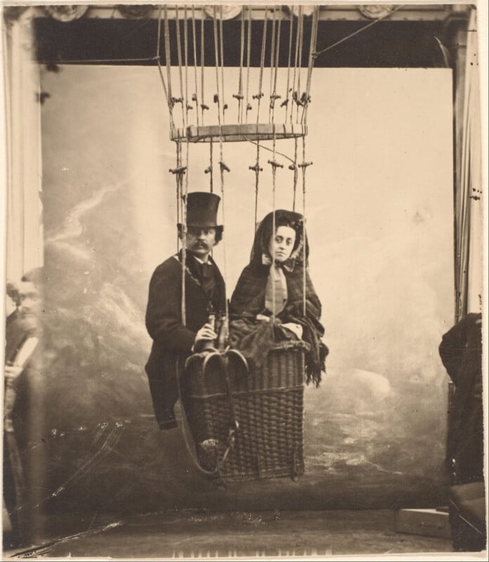 A vintage black-and-white photo shows a man in a top hat and a woman in a shawl sitting in a wicker balloon basket, suspended by ropes, with blurred figures at the sides and a painted backdrop behind them.