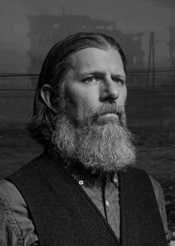 A serious, bearded man with long hair looks into the distance. He wears a button-up shirt and a vest. The background shows a dark, foggy, and ruined cityscape with silhouetted buildings and power lines. The image is black and white.