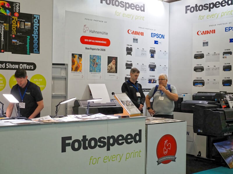 Three men stand behind a Fotospeed booth at a trade show, surrounded by printers, paper samples, and promotional posters for brands like Canon, Epson, and Hahnemühle. The booth displays the slogan “for every print.”.