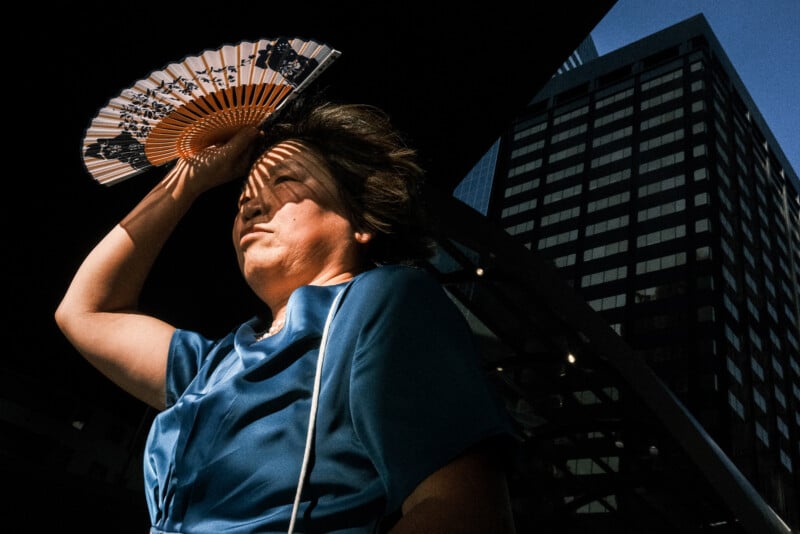 A woman in a blue dress holds a decorative hand fan above her head, casting shadows on her face, with tall modern buildings in the background under bright sunlight.