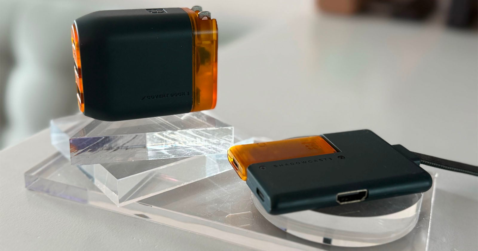 Two small electronic devices with black and translucent orange casings rest on clear acrylic stands; one device is upright, the other is flat with a visible HDMI port and the label “SHADOWCAST” on its surface.