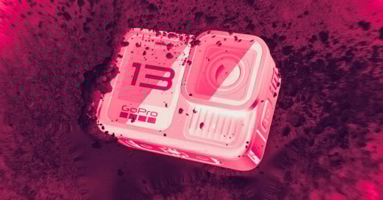 A GoPro HERO13 action camera partially covered in dirt, lying at an angle on a rough surface. The image has a bold pink and red color filter, giving it a dramatic, high-contrast appearance.