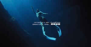 A person wearing fins and a swimsuit swims underwater in deep blue water, surrounded by rocks. Light filters down from above. Text on the image reads, "Captured on a new generation of GoPro.