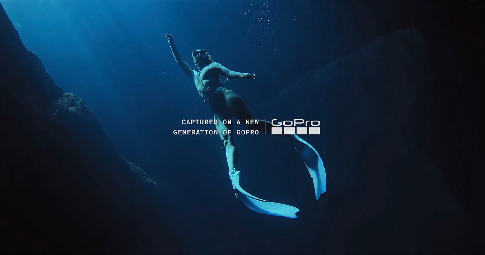 A person wearing fins and a swimsuit swims underwater in deep blue water, surrounded by rocks. Light filters down from above. Text on the image reads, "Captured on a new generation of GoPro.