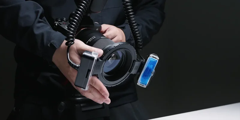 A person holding a professional camera with a macro lens, equipped with two external flash units mounted on each side and connected by coiled cables, aimed at a white surface.