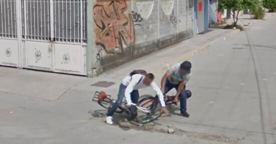 Two people are picking up their bicycles after falling into a pothole on a city street. One wears a white shirt and backpack; the other wears a hat and gray shirt. A gated building and graffiti-covered wall are visible in the background.