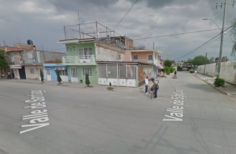 A street corner in a residential neighborhood with two-story houses, some under construction. Several people stand on the sidewalk near the intersection labeled "Valle de Santiago." The sky is overcast.