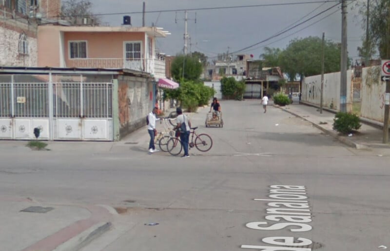 Two people stand with bicycles in the middle of a street intersection in an urban neighborhood. A man pushes a cart in the background, while another person walks further down the street. Houses and a fence line the road.