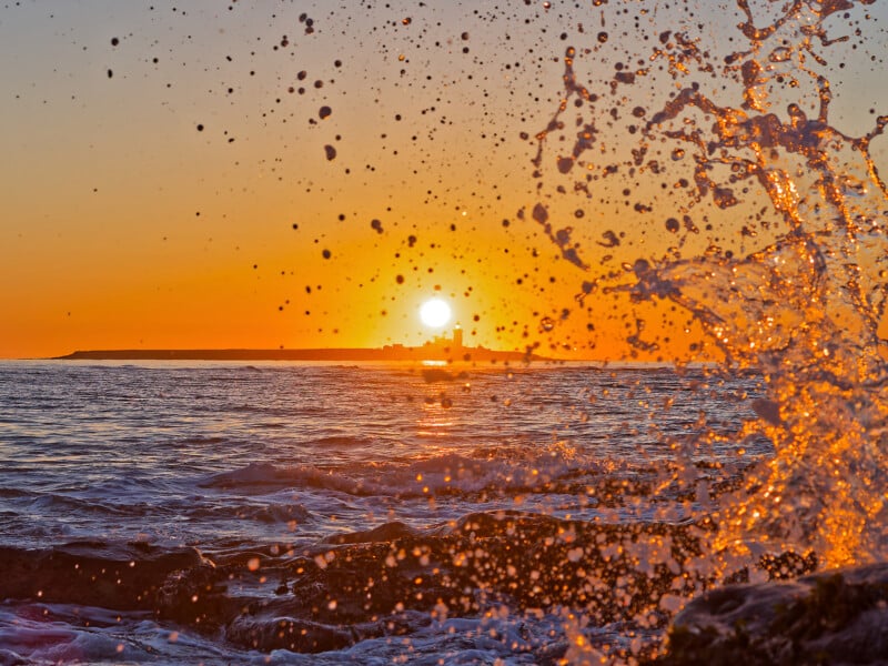 Waves splash against rocks in the foreground as the sun sets over the ocean horizon, casting an orange glow across the sky and water.