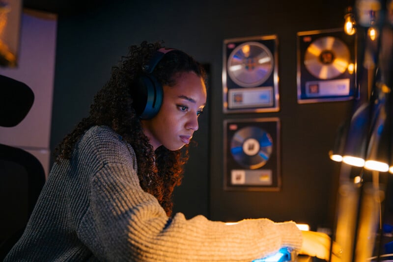 A young woman wearing headphones works at a mixing console in a dimly lit recording studio, with framed gold and platinum records displayed on the dark wall behind her.