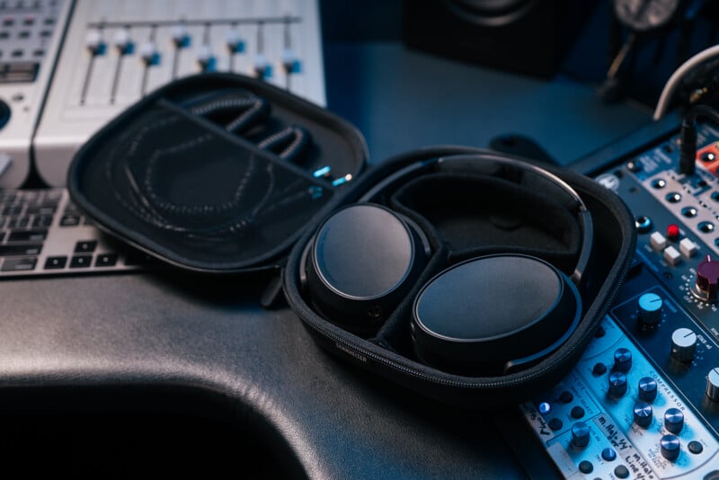 A pair of black over-ear headphones in a zippered carrying case sits on a desk in a recording studio, surrounded by audio equipment and mixing consoles.
