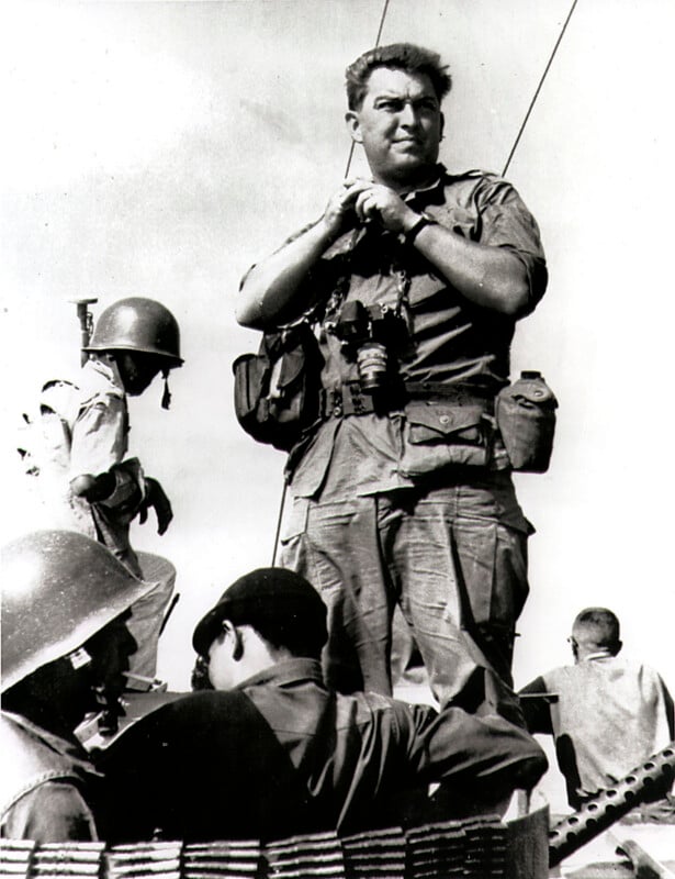 A man in military clothing stands holding binoculars with soldiers wearing helmets around him; the sky is clear in the background.