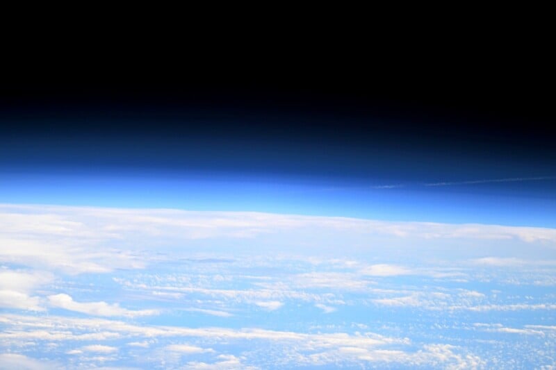 View of Earth's atmosphere from space, showing a bright blue curved horizon, scattered white clouds over the planet’s surface, and the dark blackness of outer space above.