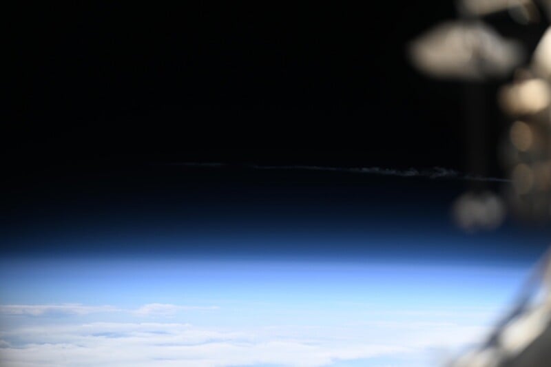 View of Earth’s atmosphere from space, showing a gradient from deep black space to blue sky above white clouds, with part of a spacecraft visible in the blurred right foreground.