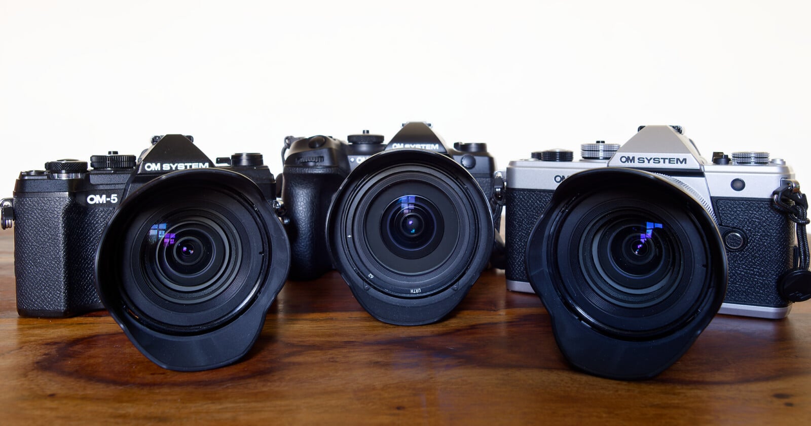 Three OM System cameras with attached lenses and hoods are displayed side by side on a wooden surface against a plain white background.